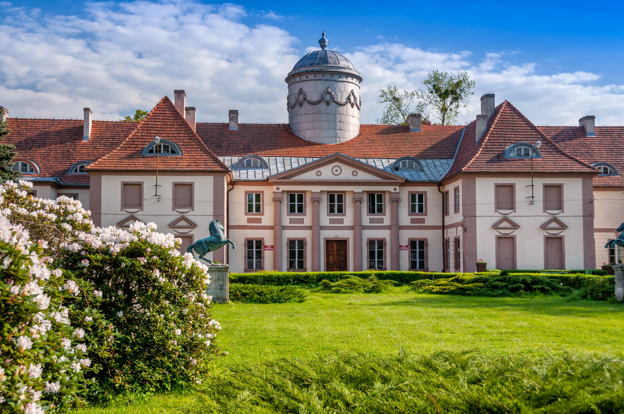 Neoklasycystyczny pałac z dużą kopułą, czerwonym dachem i rzeźbami koni w otoczeniu ogrodu